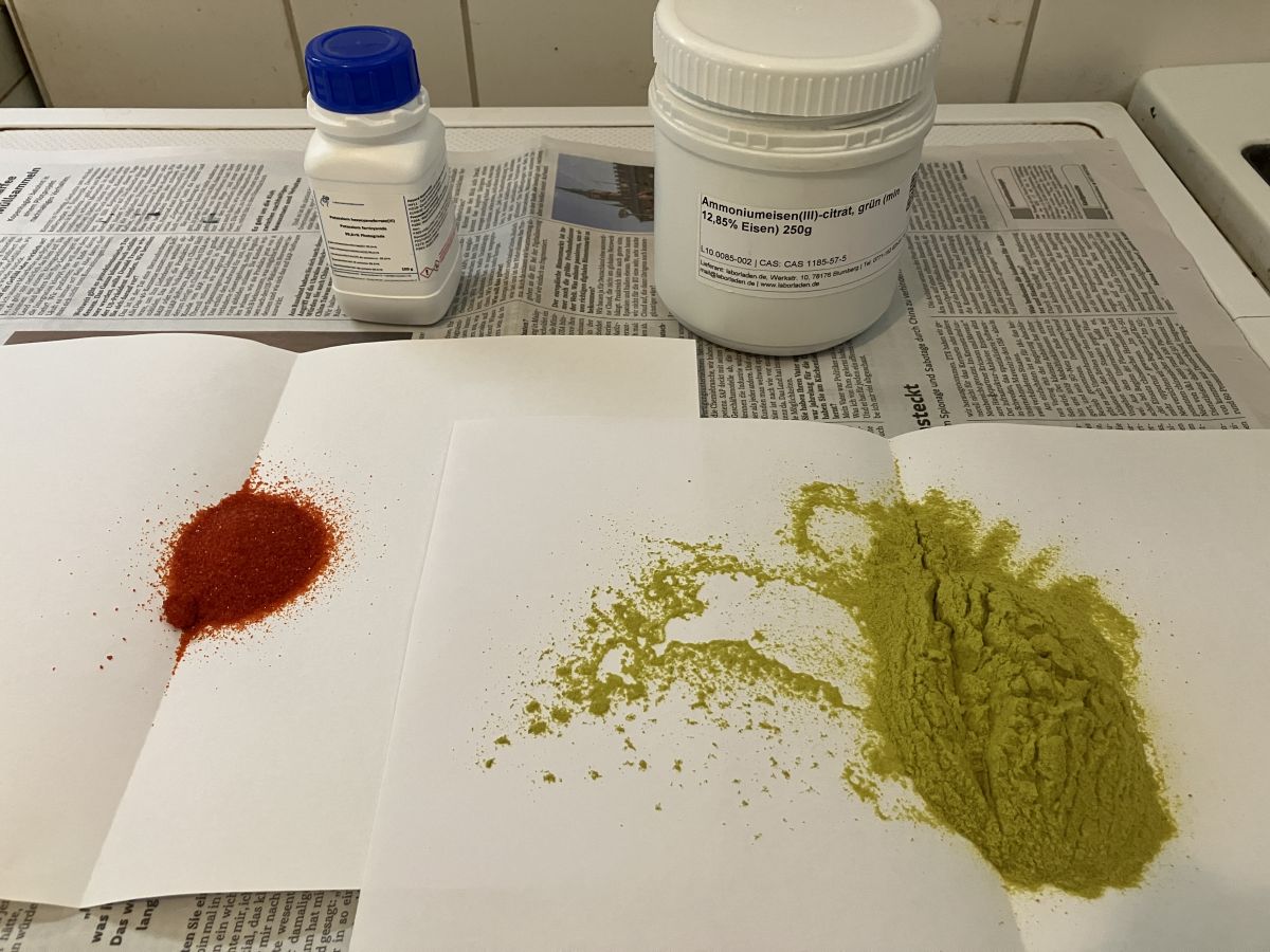 Two folded sheets of white paper. One has a small pile of red powder on it; the other has a larger pile of green powder. Two white plastic containers are in the background.