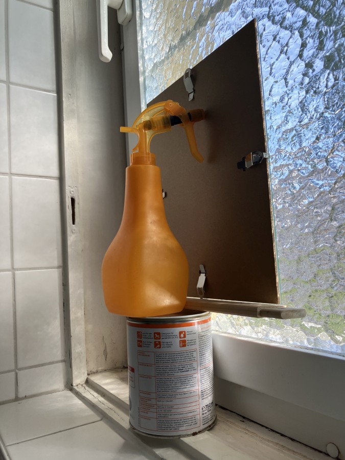 Improvised construction with a clip frame on top of a can of paint in front of a textured bathroom window. A yellow hand sprayer with water in it for weight is also on top of the can, pressing the clip frame against the window.